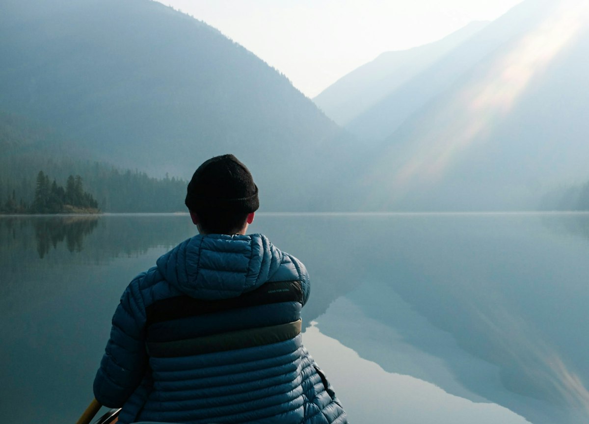 A person sitting alone in a small boat, looking out over a calm, misty lake.